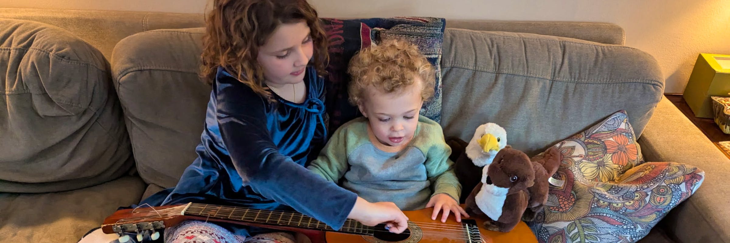 Two young kids sit on a couch with a guitar across their laps. The older child on the left shows the younger one on the right how to strum the instrument. There is a plush eagle and otter sitting next to them.