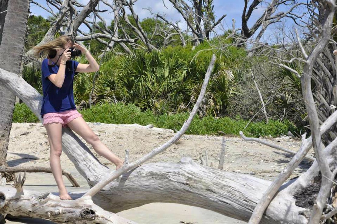 A female, Heather, sits/leans on branches of a fallen tree. She holds a camera in her hands and up to her face, photographing something off of this photo.