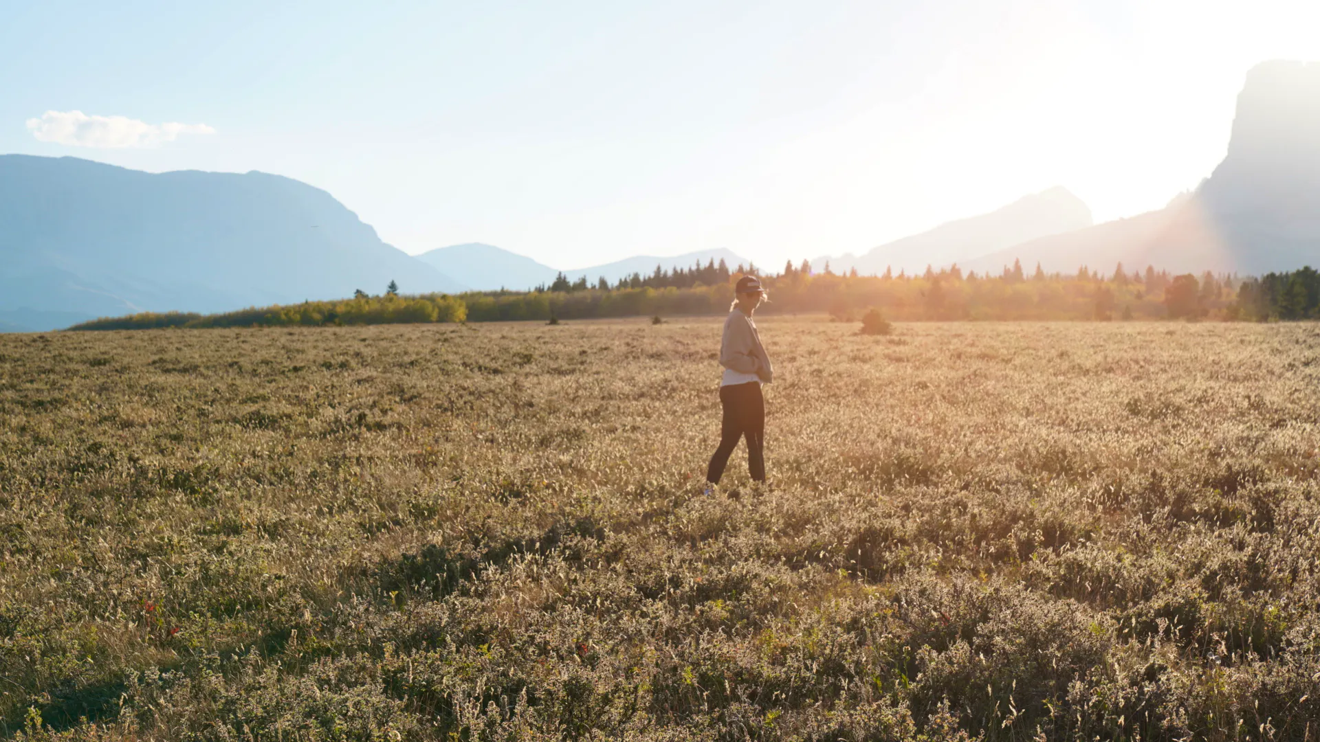 A woman walks in a field with mountains silhouetted in the distant background.