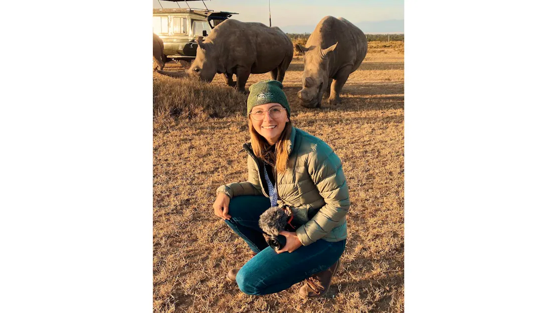Ashley Holmes crouches, holding a camera with microphones attached in her right arm. Behind her, the last two northern white rhinos graze.